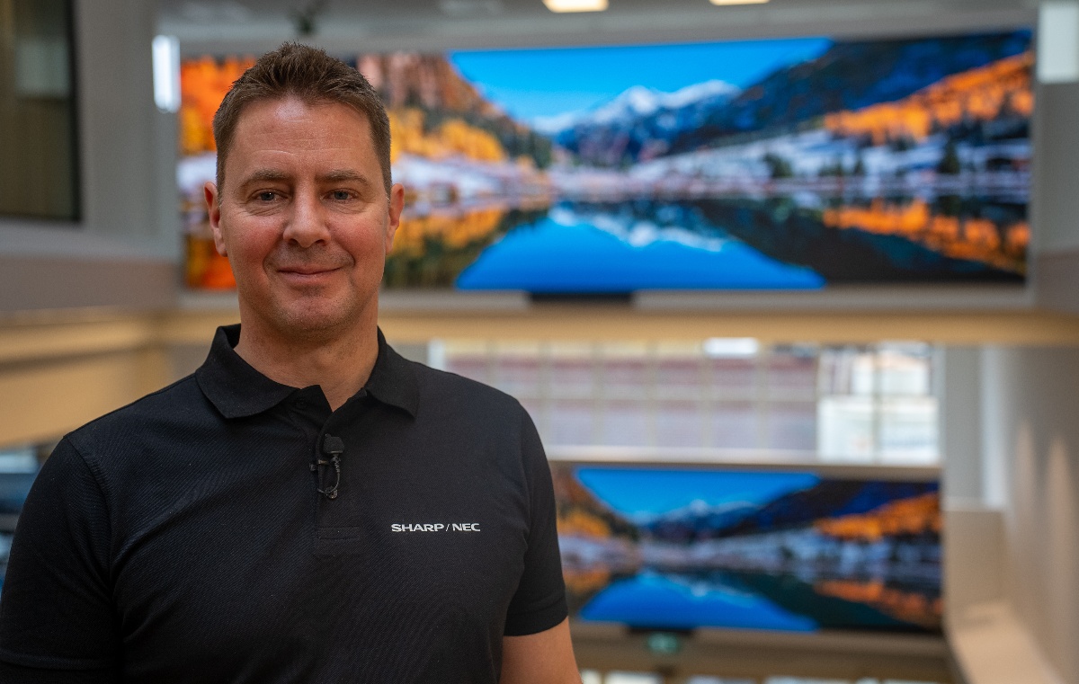 Trond Dyrnes from Sharp NEC Display Solutions standing in front of a digital screen displaying a scenic landscape of mountains and a lake.